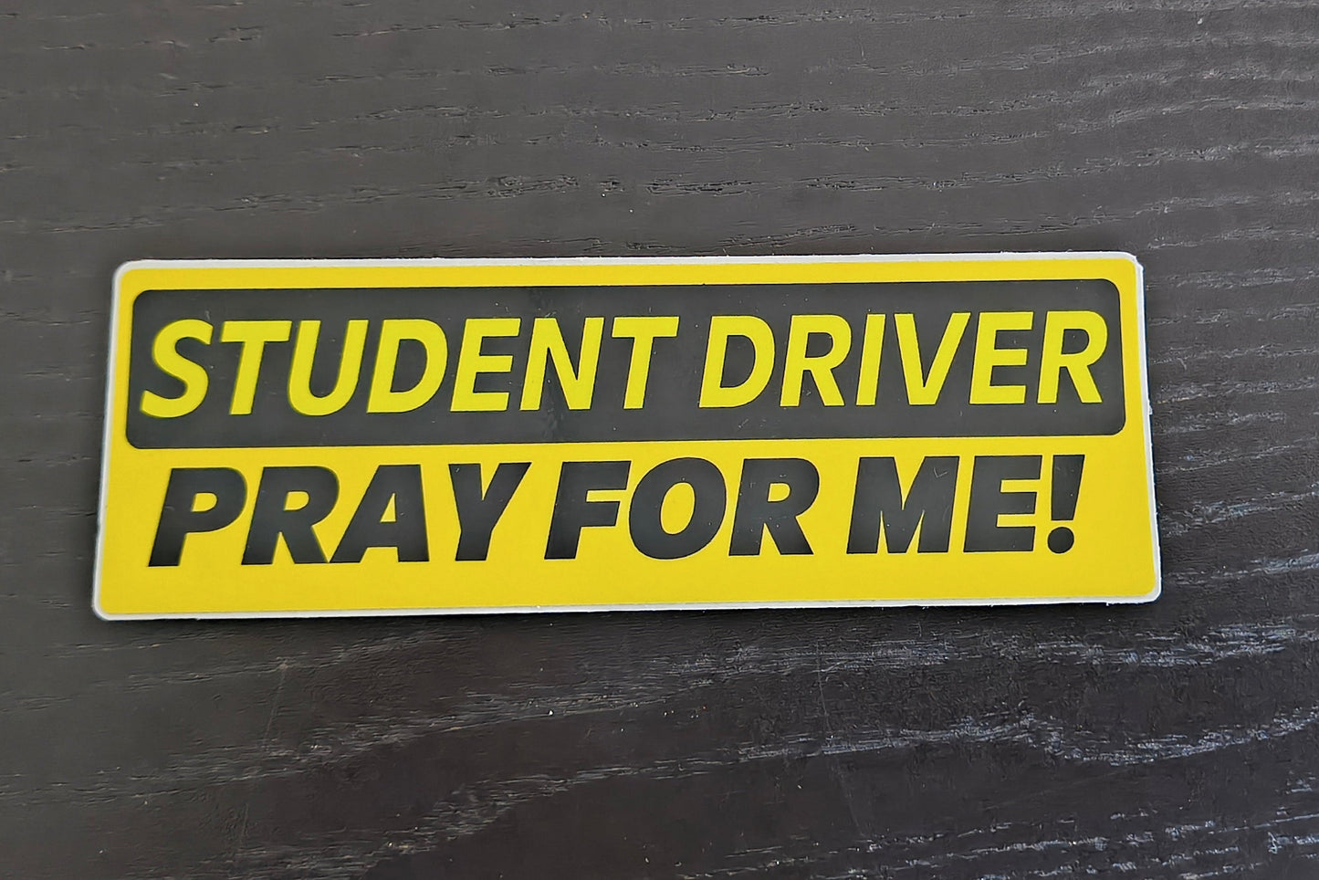 Yellow 'Student Driver Pray for Me!' sticker on a dark textured surface