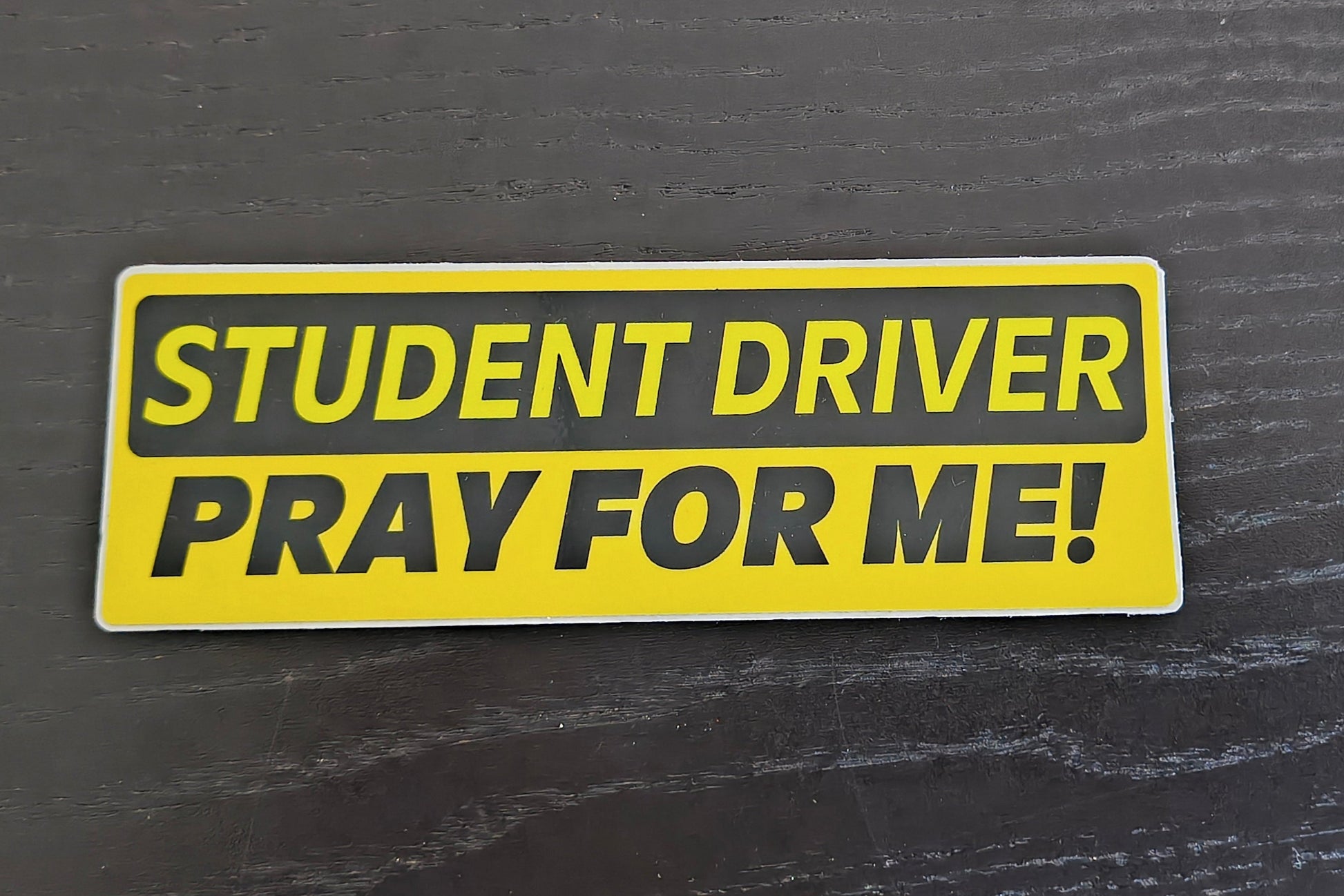 Yellow 'Student Driver Pray for Me!' sticker on a dark textured surface